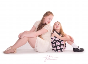 Sisters sitting on the floor back to back looking and laughing at each other. Captured by New Generation Portraits Ltd