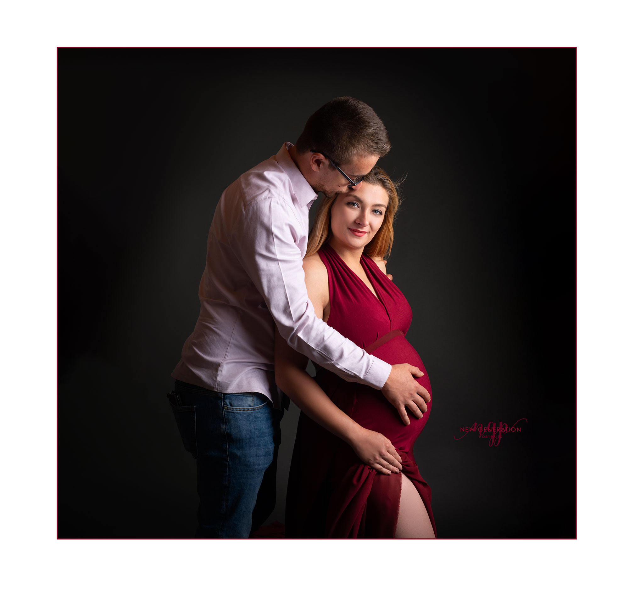 couple holding their baby bump. lady wearing red dress on dark grey background. Captured by New Generation Portraits.