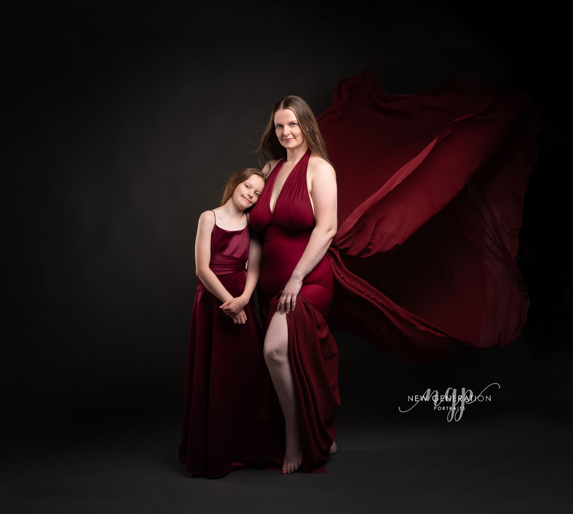 Mum and daughter on dark background wearing red dresses with wind in hair and dress. Very elegant posing. Captured by New Generation Portraits