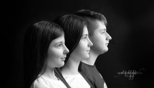 Three children all in profile in black and white on a black background. Captured by New Generation Portraits.